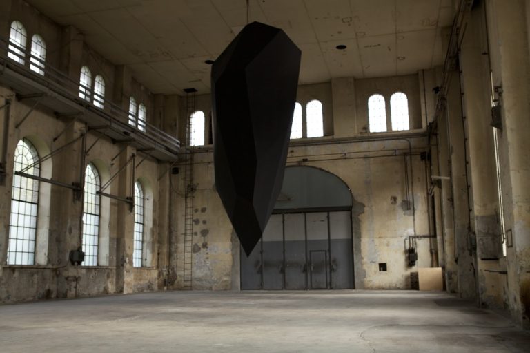 This is a photograph of an indoor space with industrial character and a black polyhedral sculpture suspended from the ceiling. The sculpture is large and appears to be made of metal or a similarly hard material.