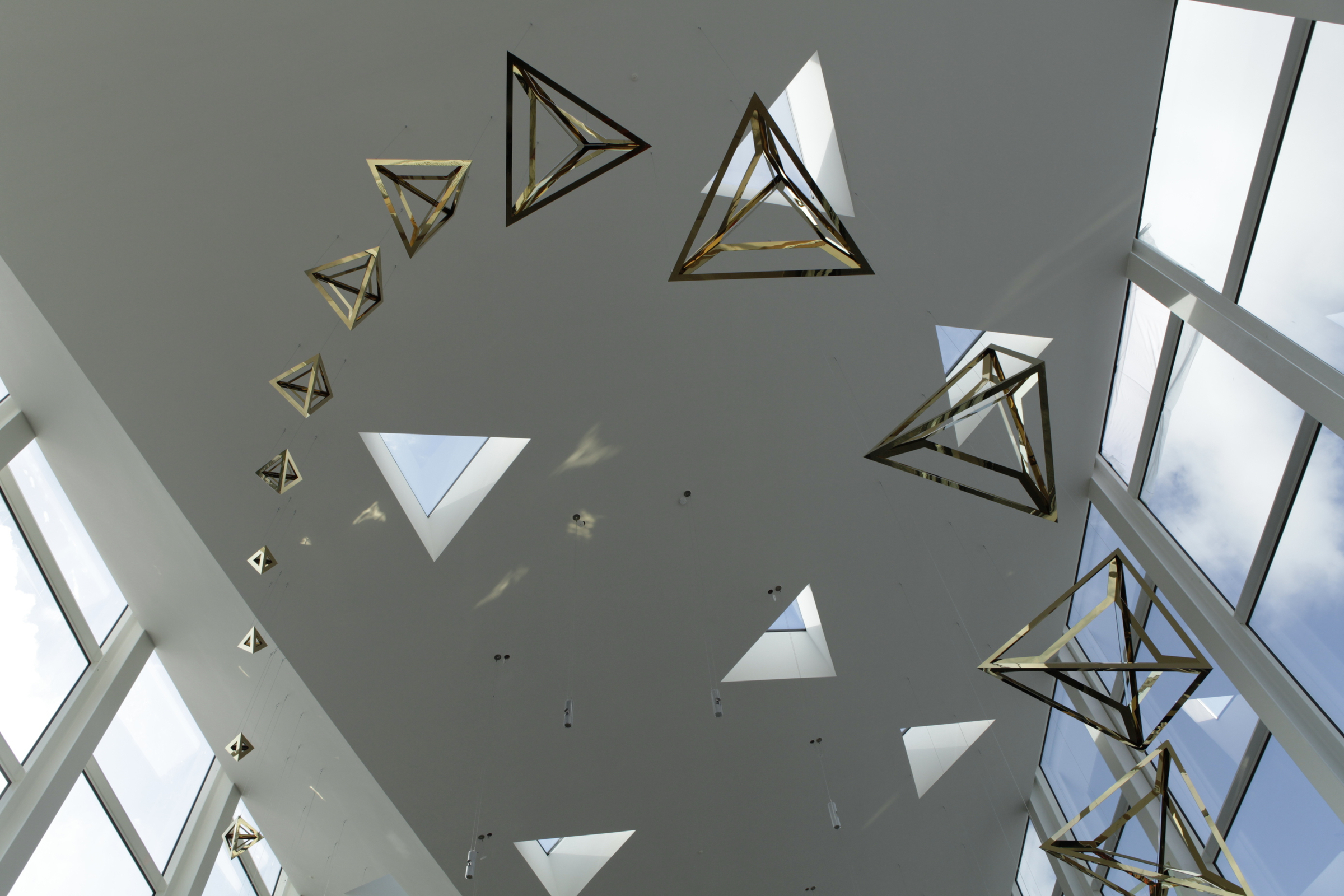 Gold-coated stainless steel tetrahedra sculpture hanging from ceiling of modern building.