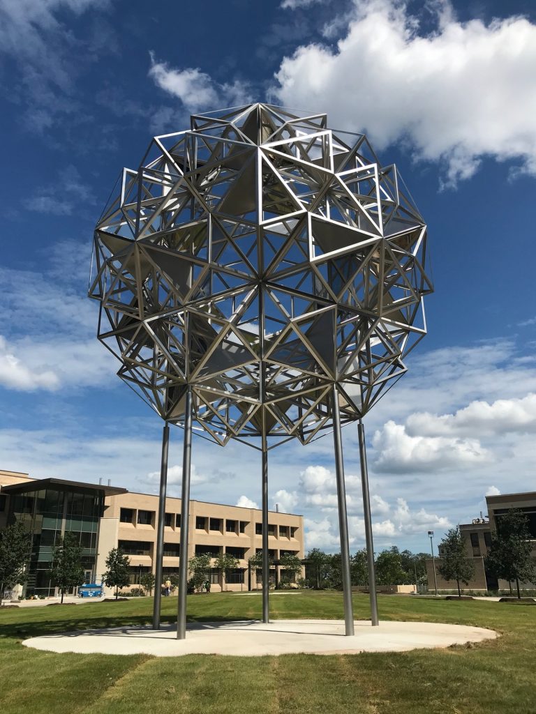 Stainless steel sculpture with projected patterns on concrete pad.