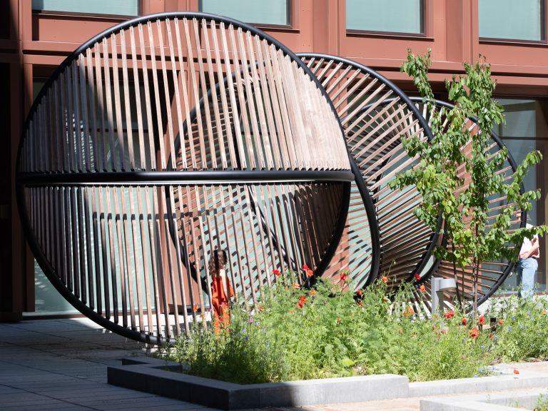 Metal sculpture featuring circular shapes and wooden texture within an outdoor courtyard setting.