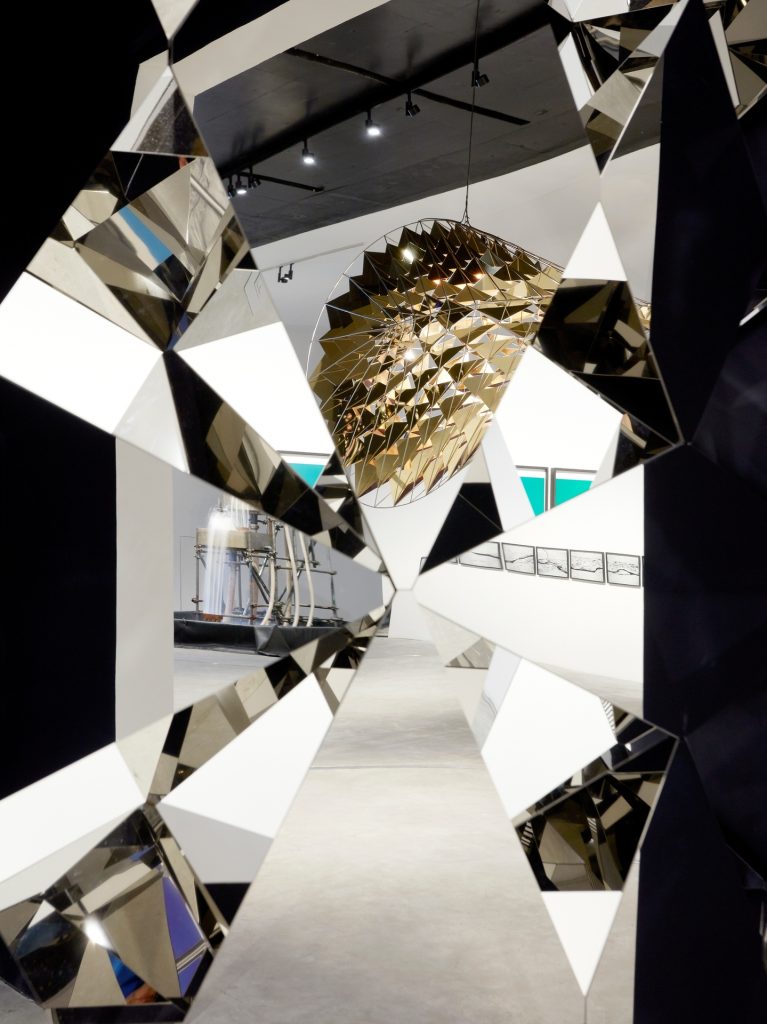 Less Ego Wall, made of stainless steel and diamond-shaped mirrors, hangs in a gallery.