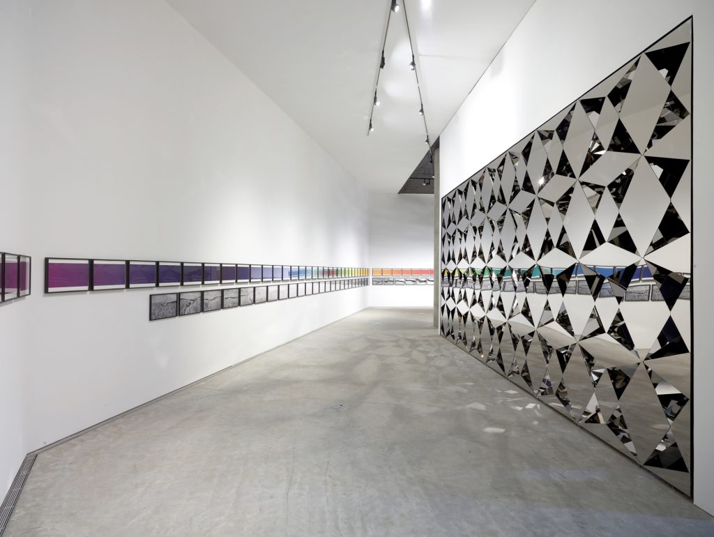 Less Ego Wall, a stainless-steel mirror-clad wall with diamond-shaped panels, is installed in an art gallery.