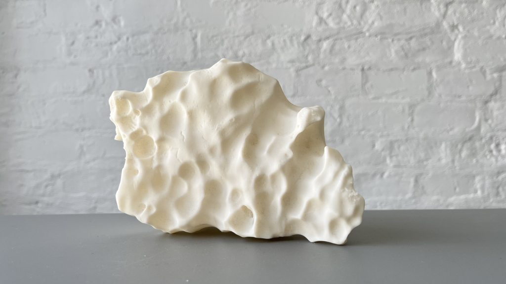 White 3D printed resin sculpture sits on gray surface against white brick wall.