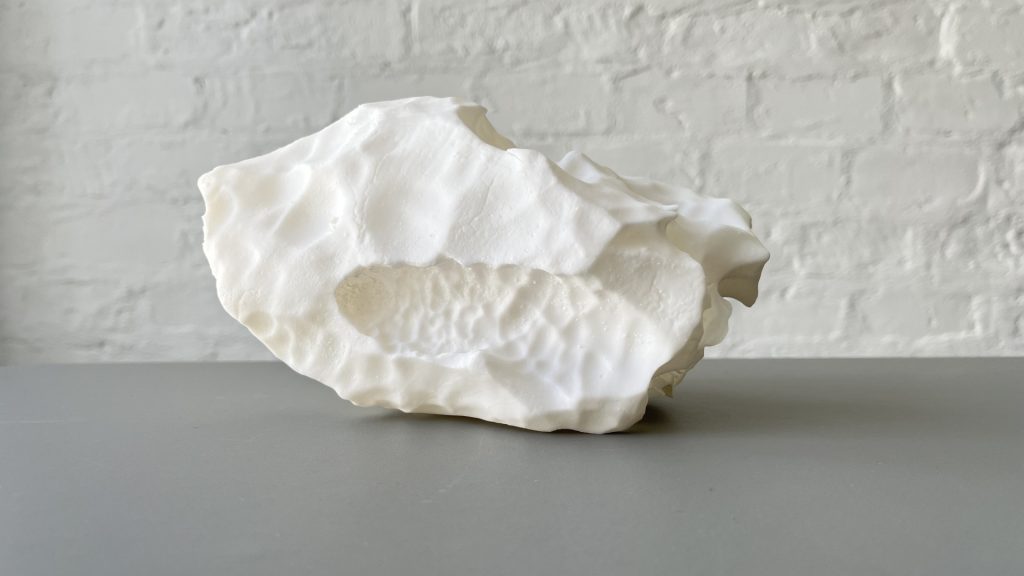White 3D-printed resin sculpture sits on gray surface against white brick wall.