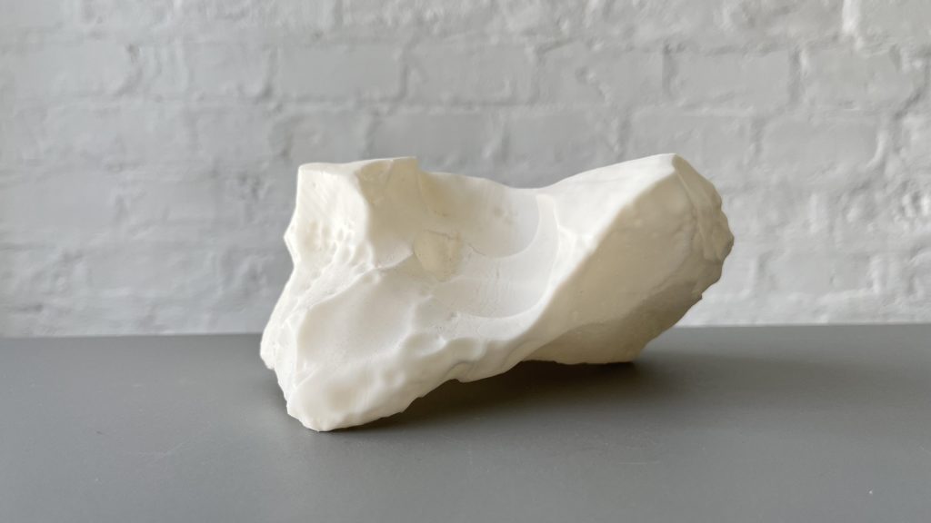White 3D-printed resin sculpture sits on gray surface against white brick wall.