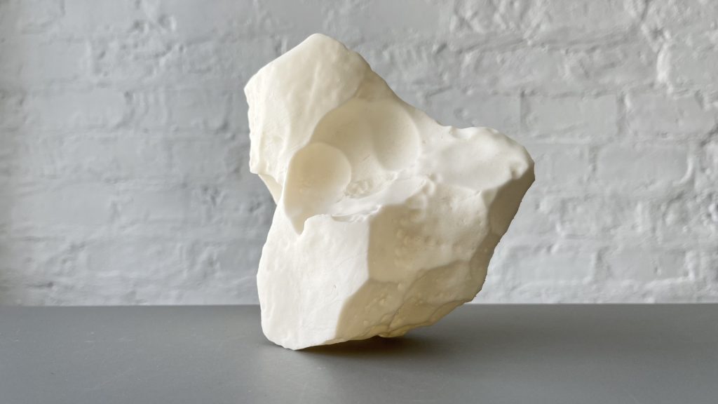 White 3D printed resin sculpture sits on gray surface against white brick wall.