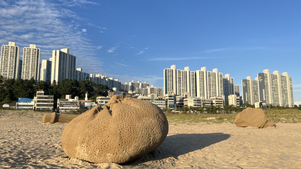 Tidal Memories" sand sculptures sit on a beach with city skyline in the background.