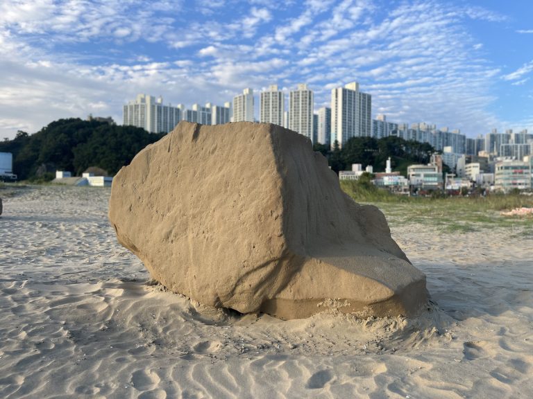 A sand sculpture sits on a beach with a city skyline in the background.