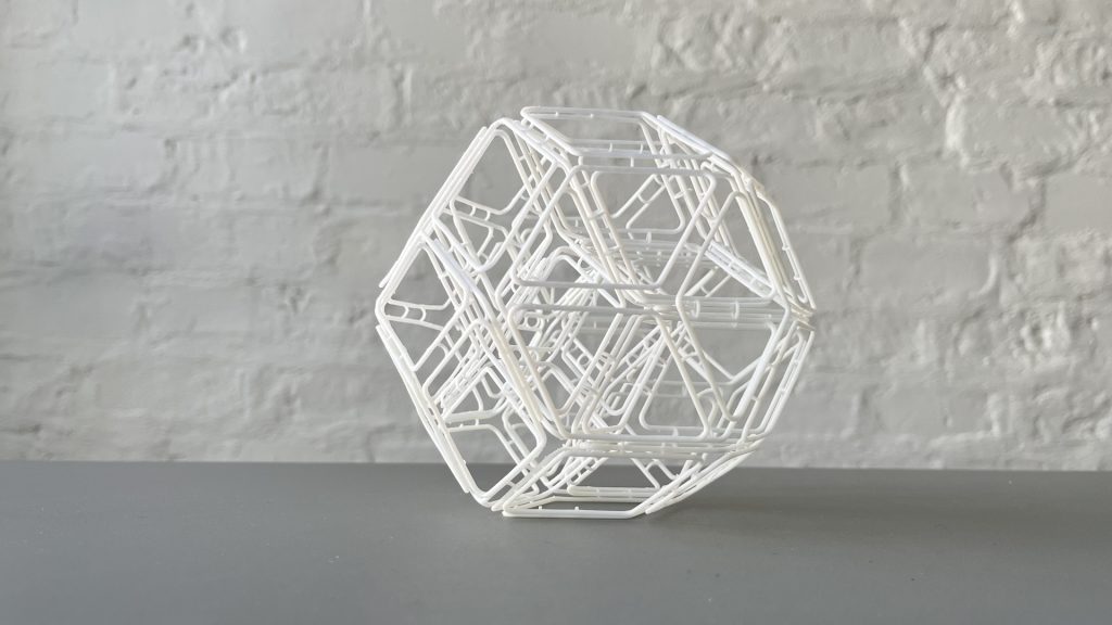 3D-printed resin rhombic structure sits on a gray surface against a white brick wall.