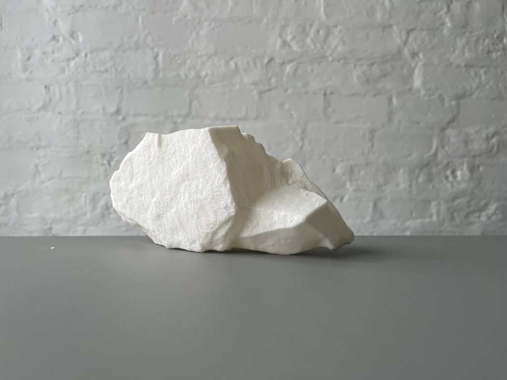 White 3D-printed resin sculpture sits on gray surface against white brick wall.