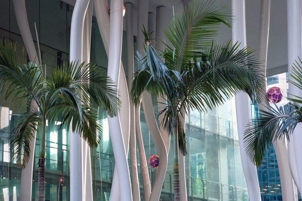 Palm trees sit among white steel columns and purple glass geometric forms in a glass-walled atrium.