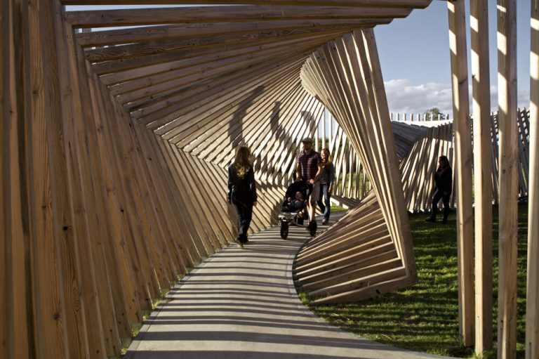 A wooden tunnel structure guides visitors through an outdoor path under sunlight.