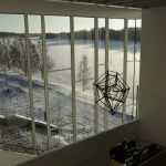 A stainless steel geometric lantern hangs from a cable near a window overlooking a snowy landscape.