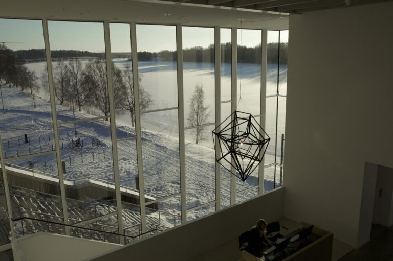 A stainless steel geometric lantern hangs from a cable near a window overlooking a snowy landscape.