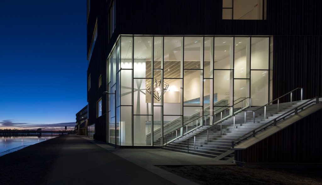 A stainless steel lantern hangs inside a glass building next to a waterfront path.
