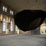 A black powder-coated polyhedron hangs from steel cables in a concrete hall with arched windows.