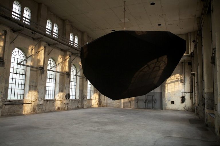 A black powder-coated polyhedron hangs from steel cables in a concrete hall with arched windows.