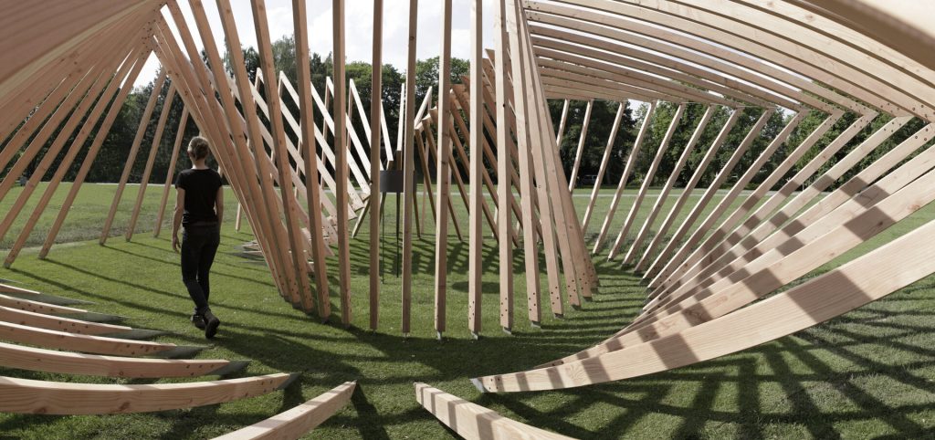 A person walks through a spiral structure of wooden slats on a grassy field.