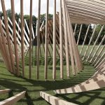 A person walks through a spiral structure of wooden slats on a grassy field.