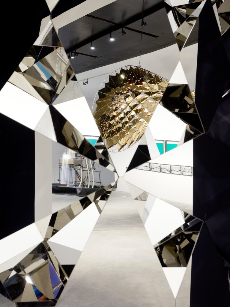 A stainless-steel mirror wall with diamond facets frames a view of a golden geometric sphere hanging in a gallery.