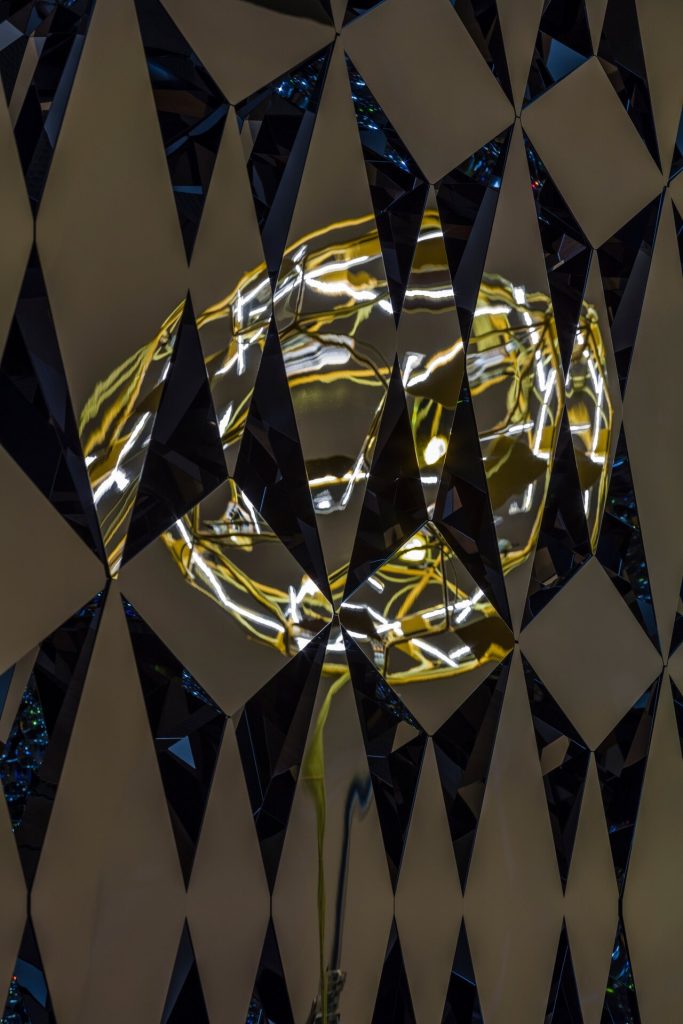 A wall of diamond-shaped stainless-steel mirrors reflects distorted images of a golden sculpture.