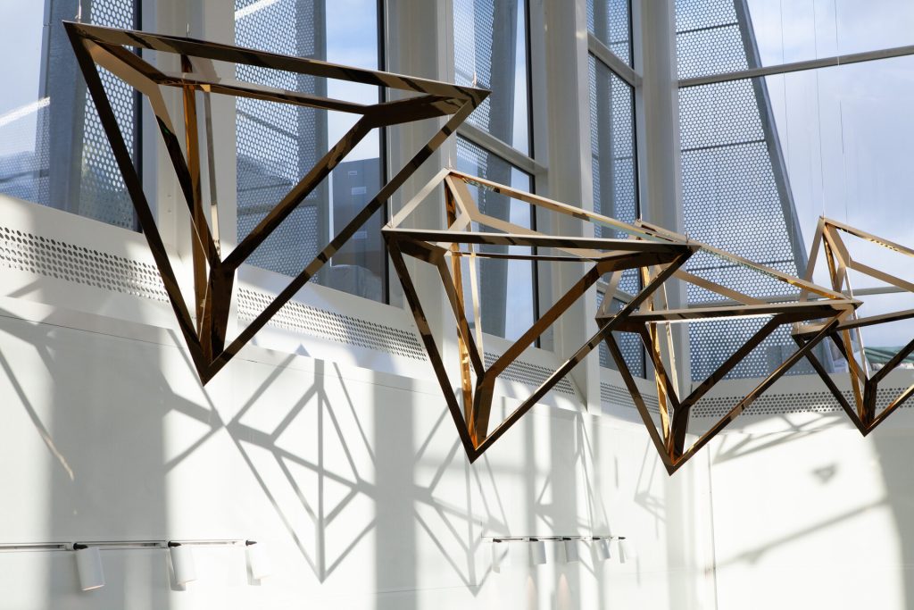 Coated stainless steel tetrahedra hang in a row, casting geometric shadows on a white wall.