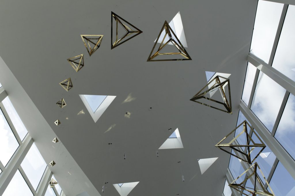 Coated stainless steel tetrahedra hang from a white ceiling with skylights and windows.