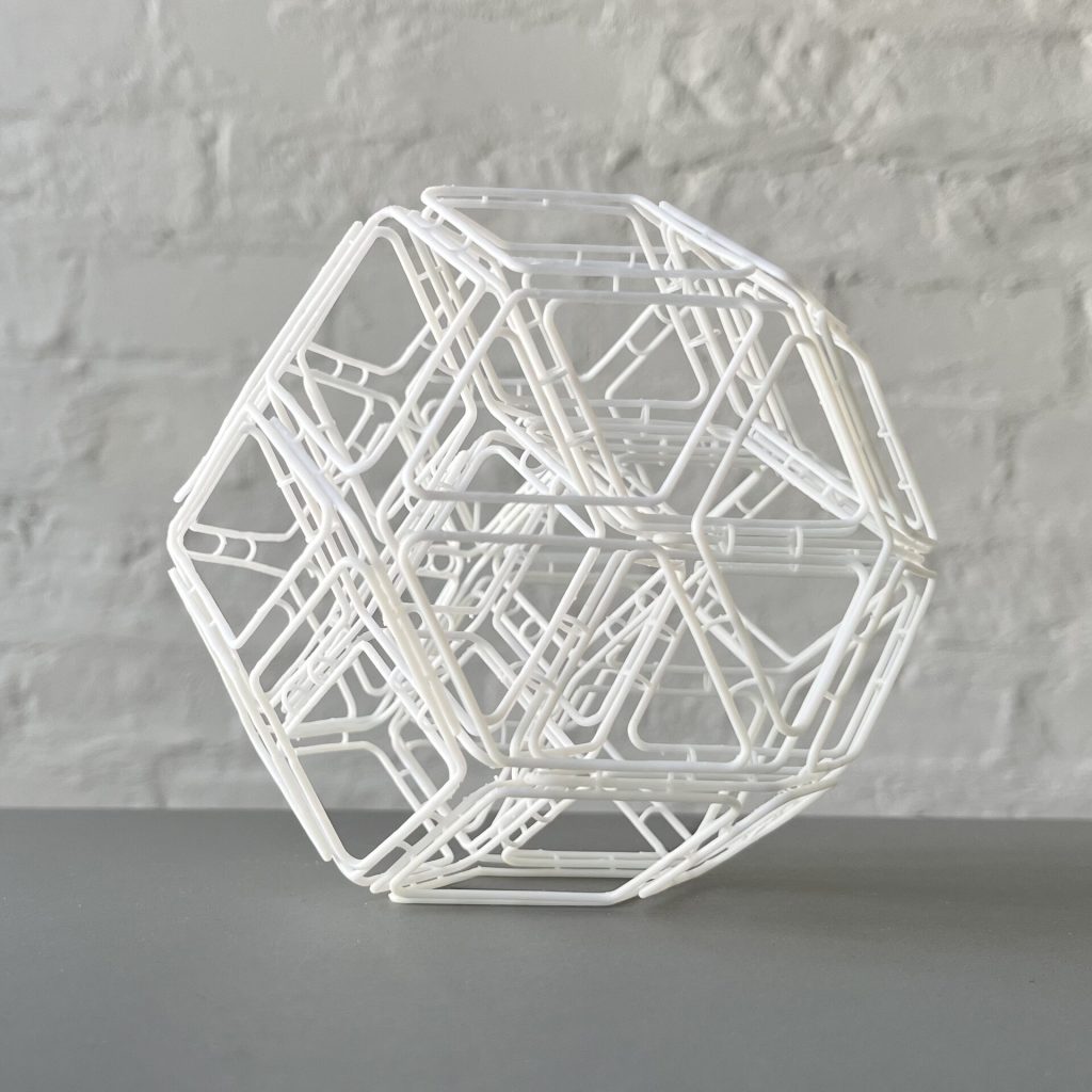 A white 3D printed resin rhombic structure sits on a gray surface against a brick wall.