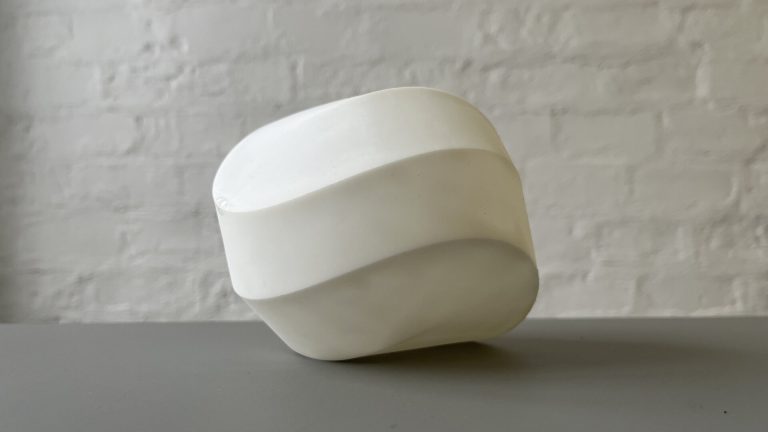 A white 3D printed resin module from the Kaleidocycloids project sits on a gray surface against a white brick wall.