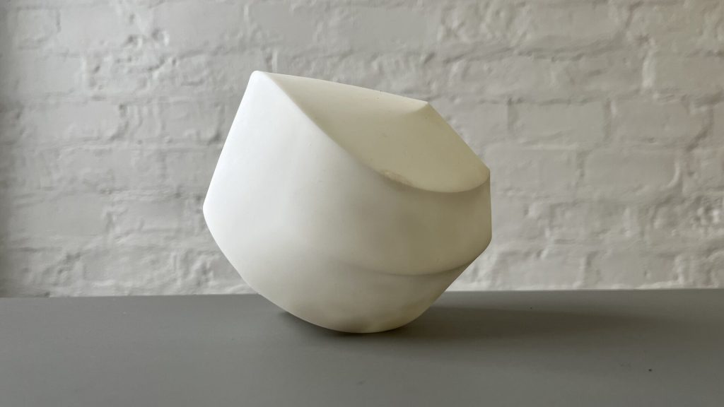 A white, 3D-printed resin object with smooth curves and angular edges sits on a gray surface against a white brick wall.