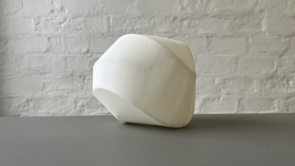 A white, 3D-printed resin object with angular facets sits on a gray surface against a brick wall.