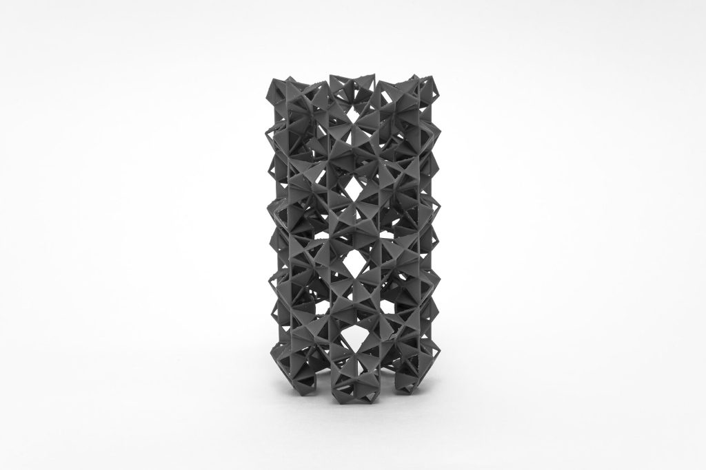 A dark gray, geometrically patterned 3D-printed resin tower from the SuperCube Towers study by Phillip C. Reiner stands vertically against a white background.