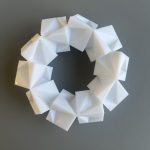 A white 3D printed resin ring of ten interlocking cube clusters sits on a gray surface.