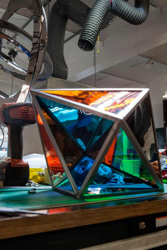 A geometric window element with colored glass panels sits on a workbench near a power drill.