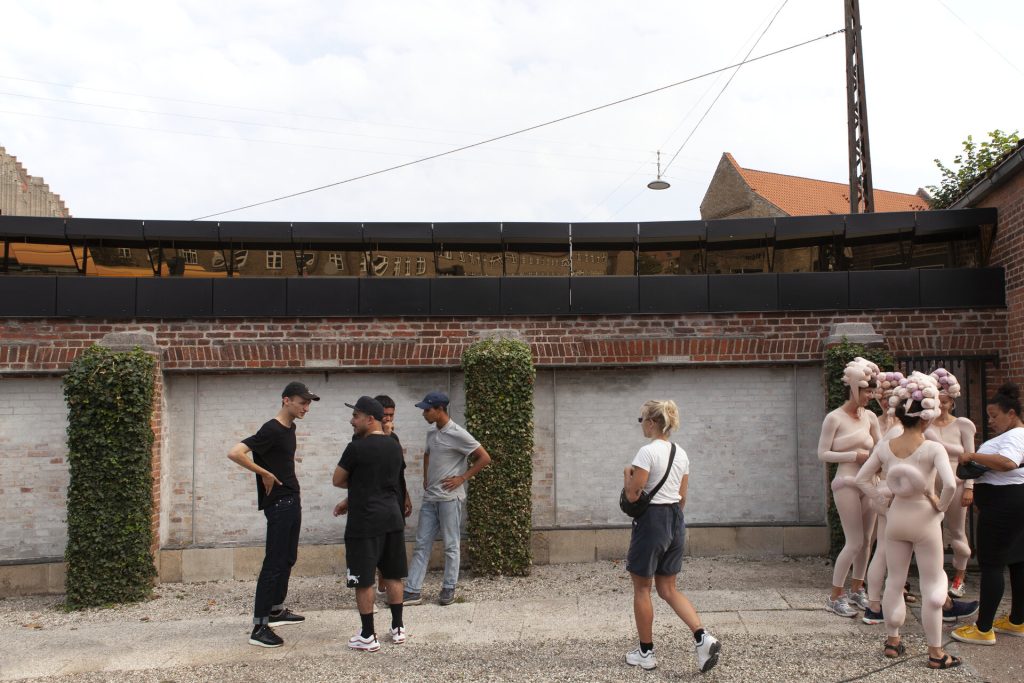 A black coated stainless steel mirror assembly hangs above a brick wall where people stand.