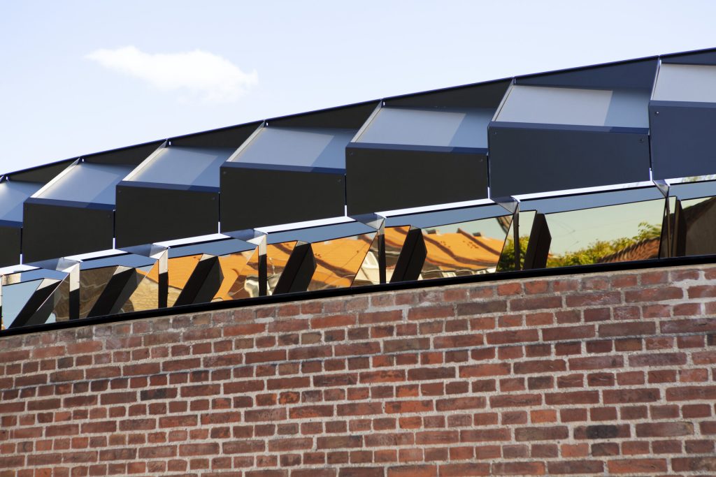 A coated stainless steel rotating mirror assembly sits on a brick wall under a blue sky.