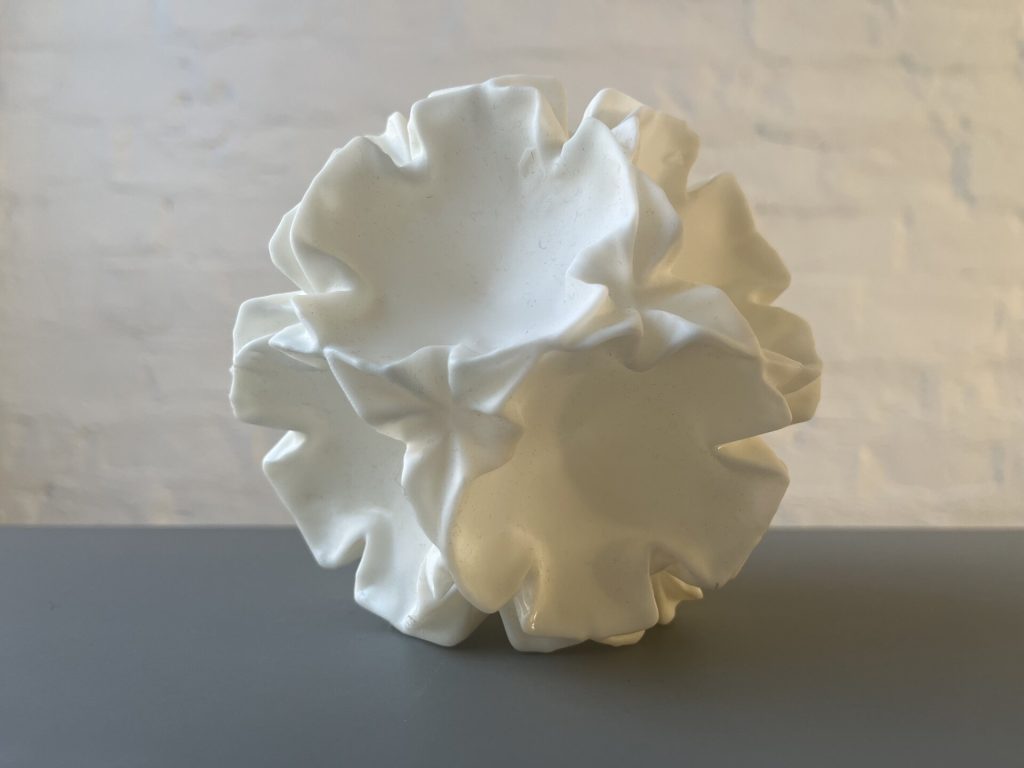 A white, 3D-printed resin sculpture resembling a crumpled flower sits on a gray surface against a light wall.