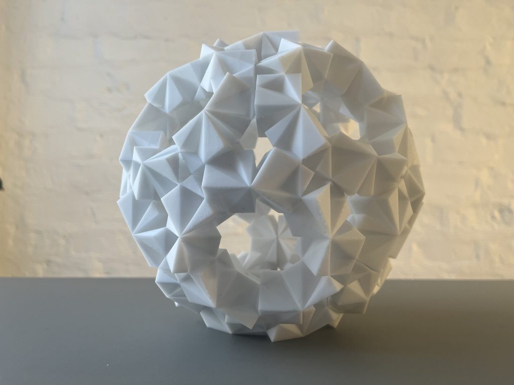 A white 3D-printed resin sculpture with triangular facets forms a spherical shape with a central opening on a gray surface.