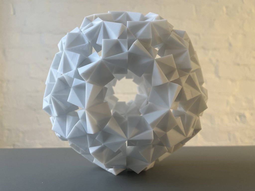 A white 3D-printed resin sculpture with interlocking triangular and pentagonal facets forms a spherical ring structure on a gray surface.