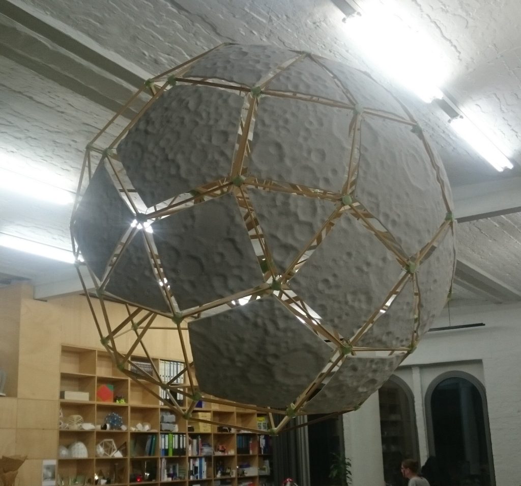 A large spherical sculpture made of 3D-printed resin panels and wooden struts hangs from the ceiling.