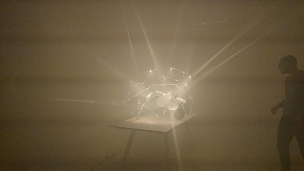 A 3D-printed resin polyhedron with parabolic mirrors emits light beams in a foggy room.