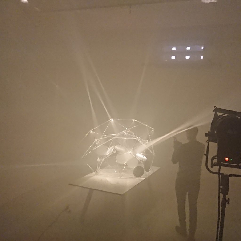 A 3D printed resin structure with parabolic mirrors sits on a table, projecting light cones in a foggy room.