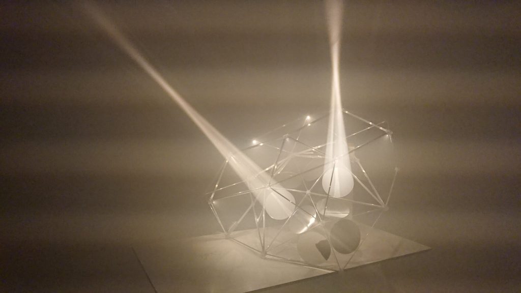 A 3D-printed resin polyhedron with internal spherical elements and light beams projects through a foggy environment.