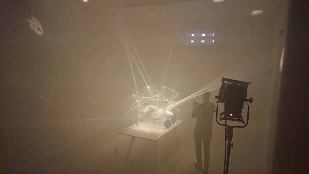 A 3D-printed resin polyhedron with internal parabolic mirrors sits on a table in a foggy room, illuminated by spotlights.