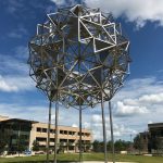 A stainless steel mirror sphere sculpture sits on four poles in a grassy area with buildings behind it under a blue sky.