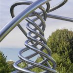 A stainless steel spiral sculpture sits outdoors with trees and sky in the background.