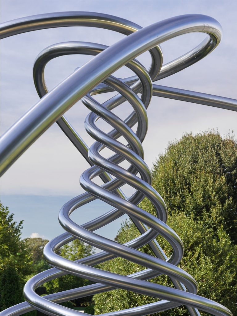 A stainless steel spiral sculpture sits outdoors with trees and sky in the background.