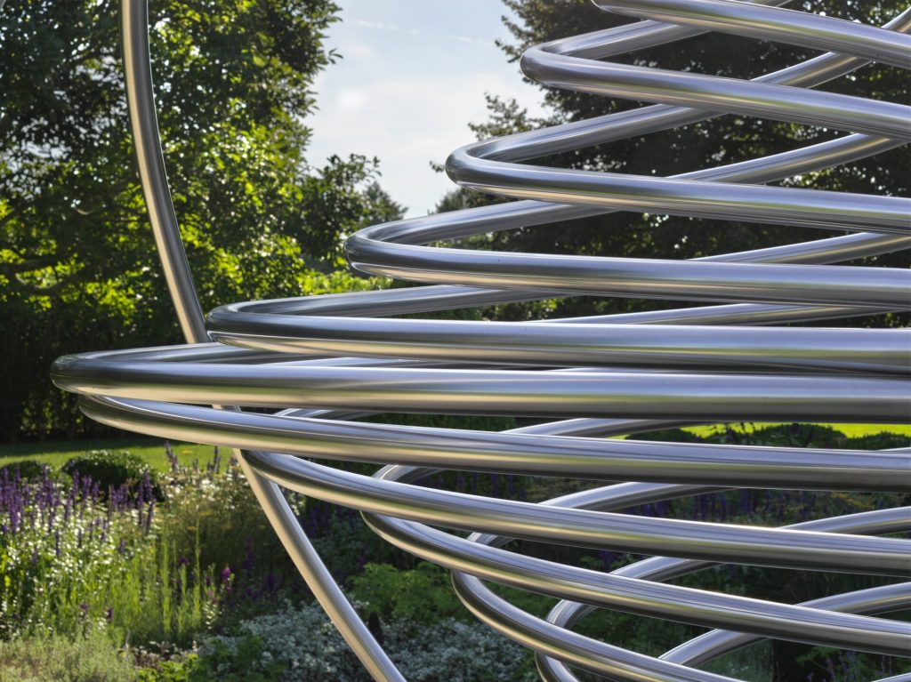 A stainless steel spiral sculpture sits in a garden with green trees and purple flowers.
