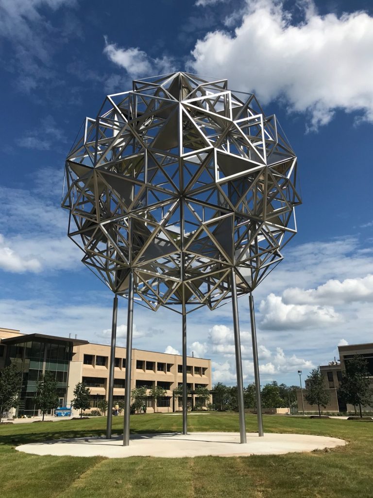 A stainless steel mirrored sphere sculpture sits on four poles in a grassy area near buildings under a blue sky with clouds.