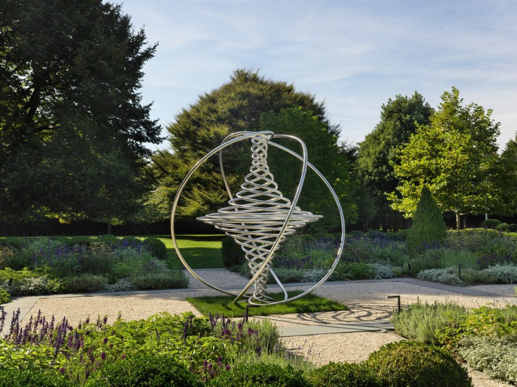 A stainless steel spiral sculpture sits on a grassy patch surrounded by gravel paths and flowering plants in a garden setting.